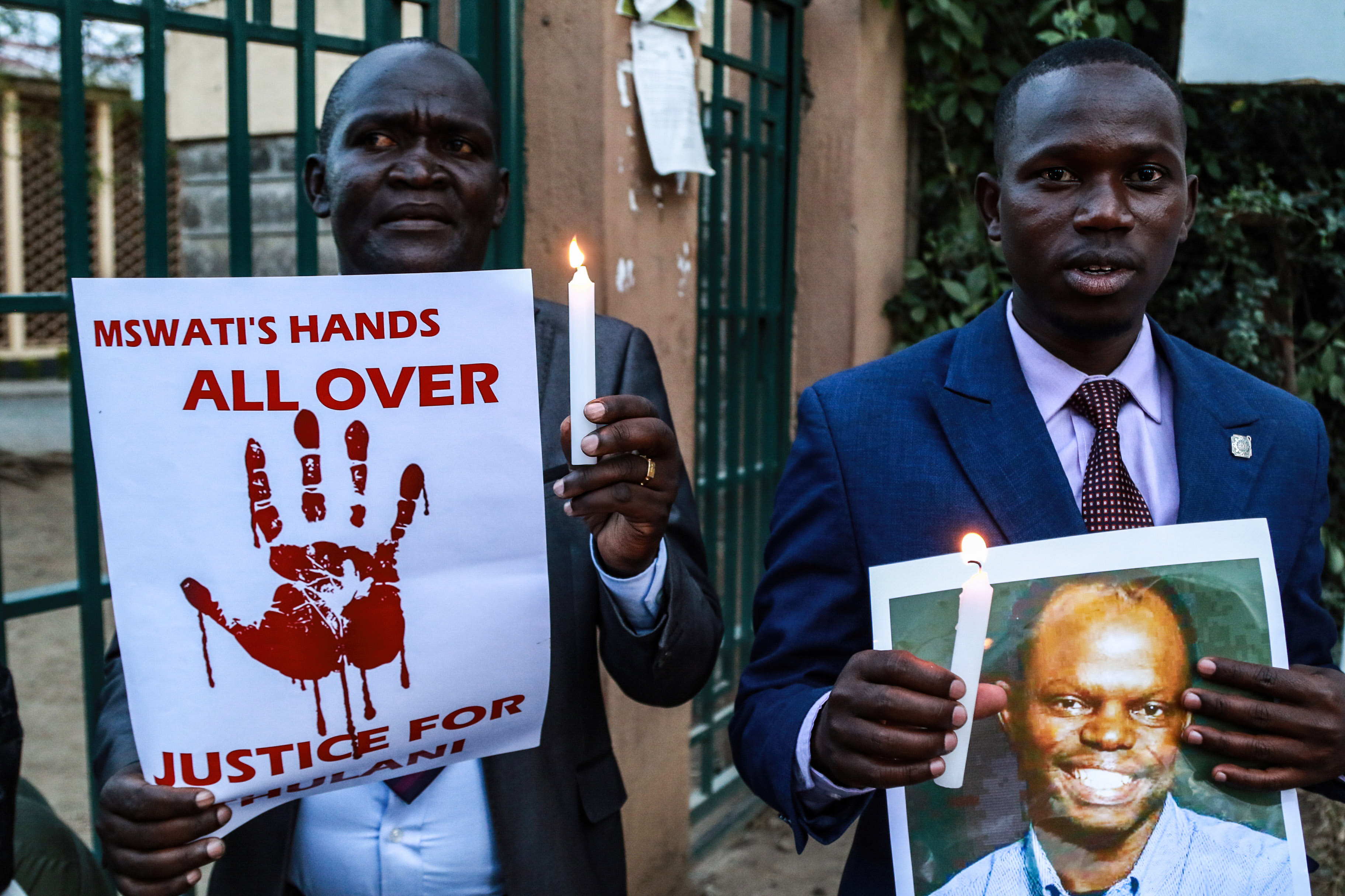 Two men stand in front of a gate holding candles, one holding a signs with a bloody hand print, the other holding a photo of a smiling man.