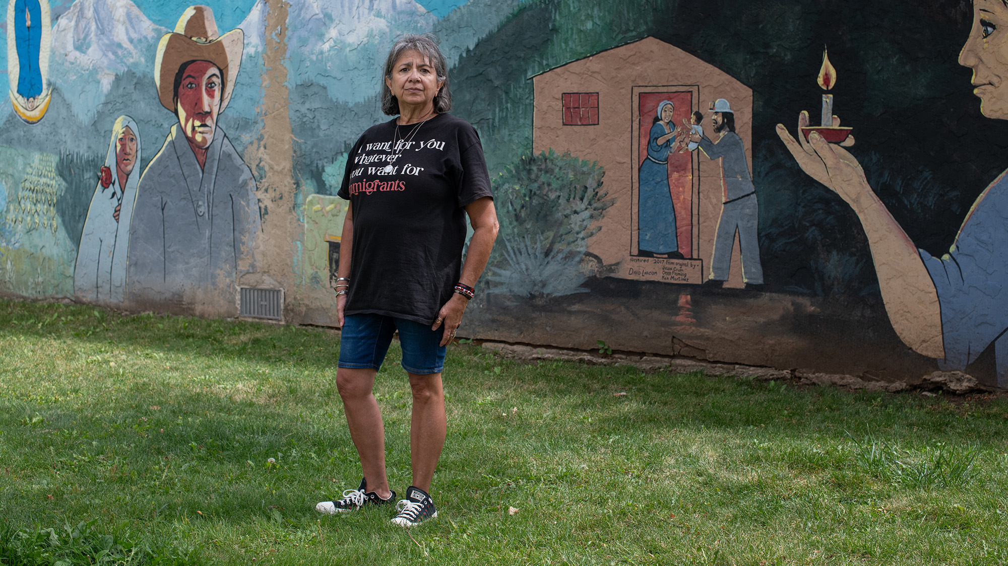 Dee Maes-Sandoval, dressed in shorts and a T-shirt, stands in front of a painted mural. The mural depicts a woman in a white shawl holding a red rose, a man wearing a beige cowboy hat, and a house where a man hands a baby to a woman standing in the doorway.