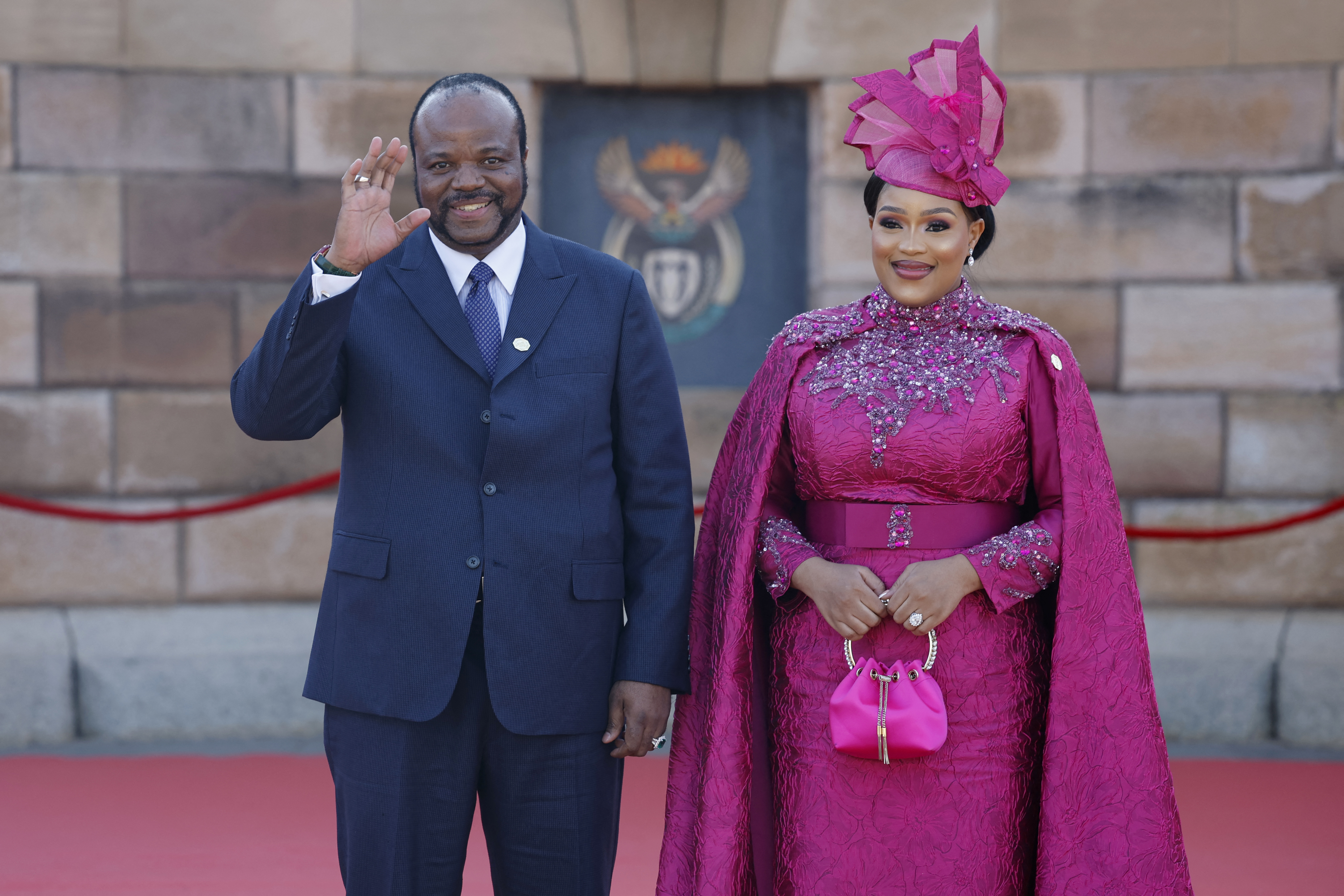 Smiling man in suit, waving, with a smiling woman dressed in pink standing next to him.